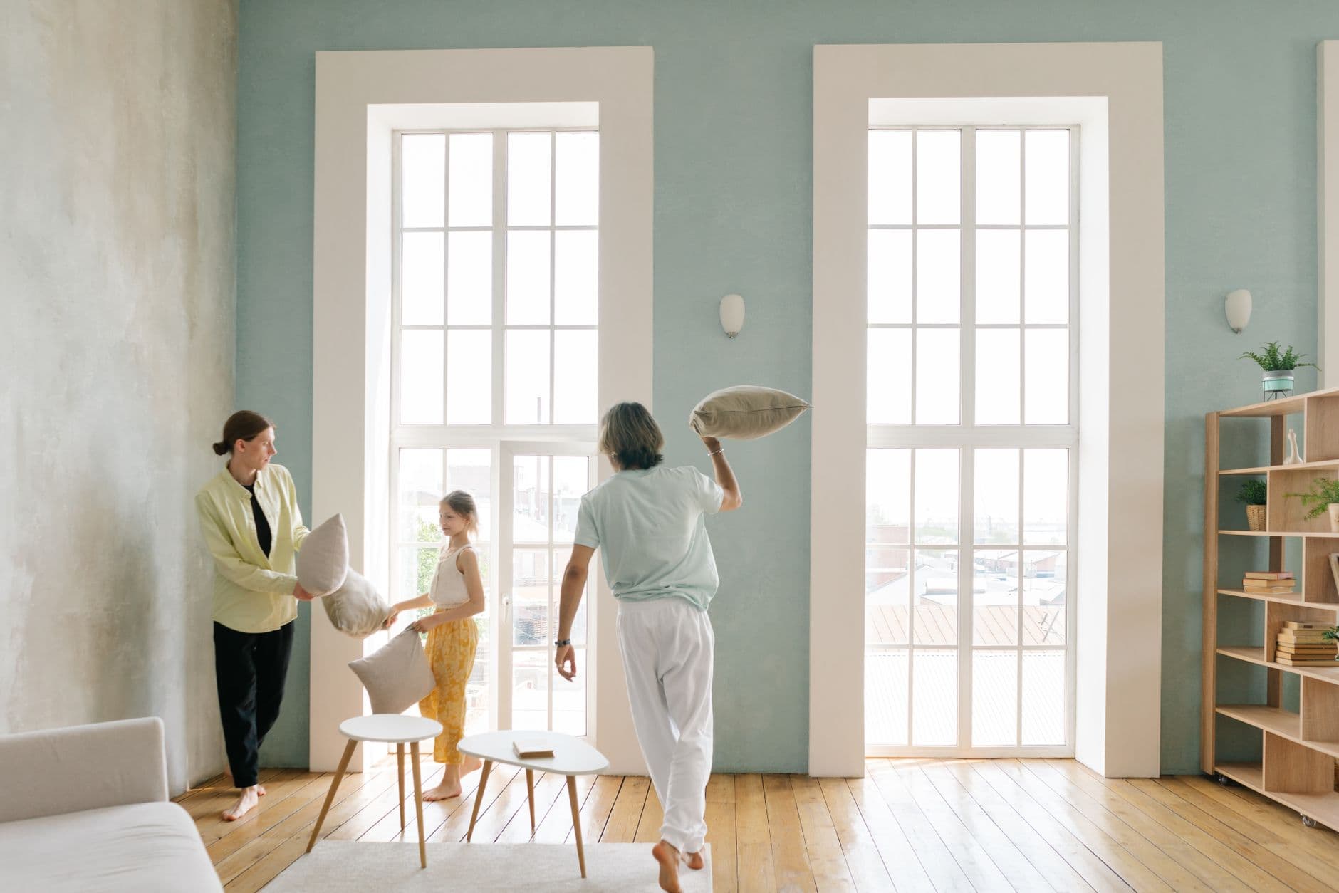 Playful family pillow fight in a bright modern living room with large windows.